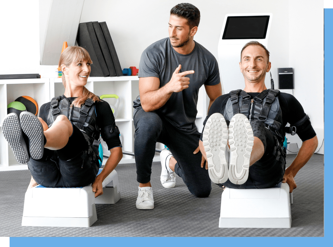 Trainer coaches two people wearing EMS suits as they perform leg raises on white exercise benches in a gym setting, smiling.